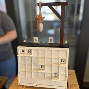 Puede incluir: Un juego de novedad con una horca de madera y una figura de vaquero colgando de ella. El juego tiene una cuadrícula blanca con letras que deletrean "ABARZ". El juego está sobre una caja de madera.