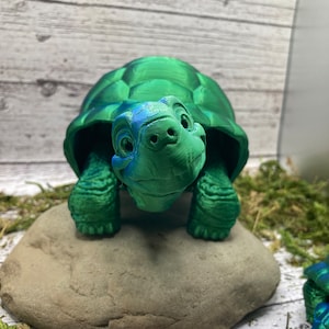 Könnte beinhalten: Eine grüne 3D-gedruckte Schildkrötenfigur mit einer leicht geöffneten Schale. Die Schildkröte sitzt auf einem grauen Stein.