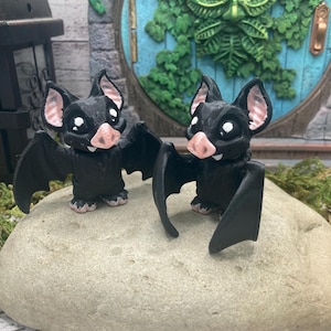 Puede incluir: Dos figuritas de murciélago negras con narices rosas y ojos blancos. Los murciélagos están posados con las alas extendidas. Están sentados en una roca gris.