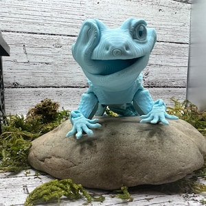 Könnte beinhalten: Hellblaue, 3D-gedruckte Eidechsenfigur mit einem lächelnden Gesichtsausdruck. Die Eidechse sitzt auf einem grauen Stein, umgeben von grünem Moos. Die Figur hat detaillierte Texturen und steht vor einem weißen Holzhintergrund.