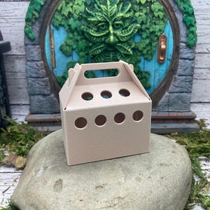 Może przedstawiać: Małe, beżowe pudełko kartonowe z uchwytem i sześcioma okrągłymi otworami. Pudełko stoi na dużym, szarym kamieniu.
