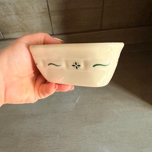 Op de afbeelding: Een crèmekleurige keramische kom met een horizontale band met een herhalend reliëfpatroon in de vorm van een raster. De kom is versierd met kleine, donkergroen geschilderde accenten, waaronder een centraal bloemmotief met vier blaadjes.