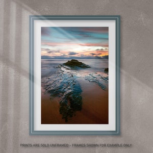 May include: Framed print of a coastal landscape featuring a rock formation in the ocean at sunset. The sky displays hues of blue, orange, and pink. The print is in a teal frame and the text at the bottom reads "PRINTS ARE SOLD UNFRAMED - FRAMES SHOWN FOR EXAMPLE ONLY".