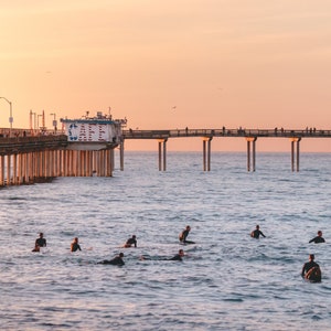 May include: A scenic view of a pier extending into the ocean at sunset. Surfers are in the water, waiting for waves. The sky is a warm blend of orange and pink.