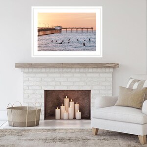 May include: A framed photograph of a pier extending into the ocean at sunset. Surfers are in the water. The sky is a gradient of orange and pink. The photograph is displayed above a fireplace with candles and a basket.