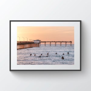 May include: A framed photograph of a pier extending into the ocean at sunset. Surfers are in the water, and the sky is a gradient of orange and pink. The pier has a building at the end and is lined with people.