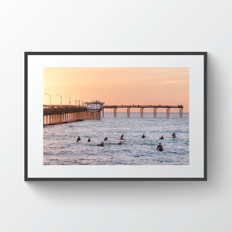 May include: A framed photograph of a pier extending into the ocean at sunset. Surfers are in the water, and the sky is a gradient of orange and pink. The pier has a building at the end and is lined with people.