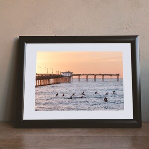 May include: A framed photograph of a pier extending into the ocean at sunset. Surfers are in the water, and the sky is a gradient of orange and pink. The frame is black, and the photograph is on a wooden surface.