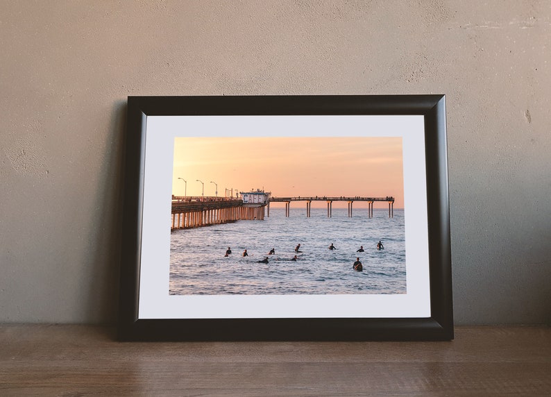 May include: A framed photograph of a pier extending into the ocean at sunset. Surfers are in the water, and the sky is a gradient of orange and pink. The frame is black, and the photograph is on a wooden surface.