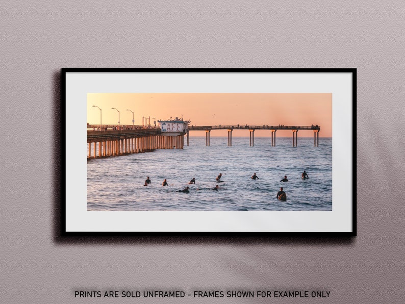 May include: A framed print of a pier extending into the ocean at sunset. Surfers are in the water, and people are on the pier. The sky is a gradient of orange and pink. The text at the bottom reads "PRINTS ARE SOLD UNFRAMED - FRAMES SHOWN FOR EXAMPLE ONLY."