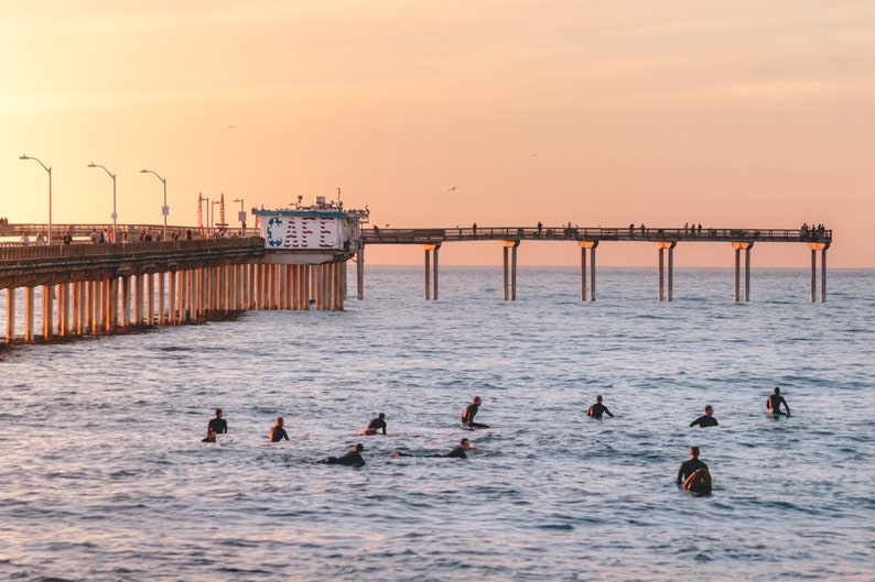 May include: A scenic view of a pier extending into the ocean at sunset. Surfers are in the water, waiting for waves. The sky is a warm blend of orange and pink.