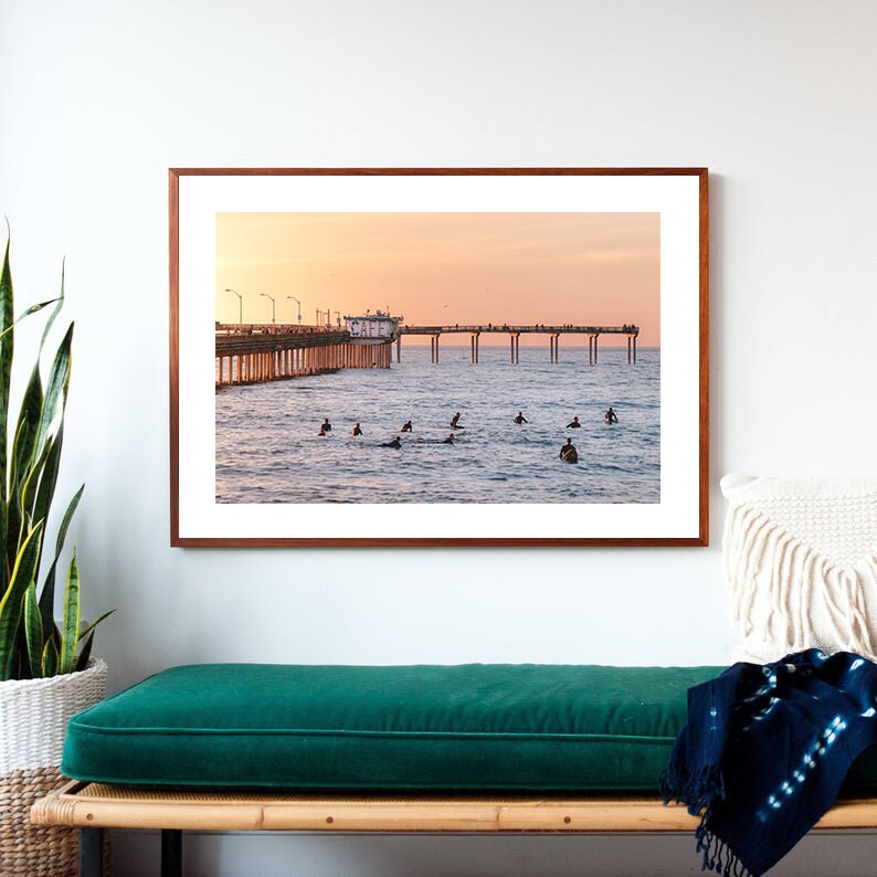 May include: A framed photograph of a pier extending into the ocean at sunset. Surfers are in the water, and people are on the pier. The sky is a gradient of orange and pink.