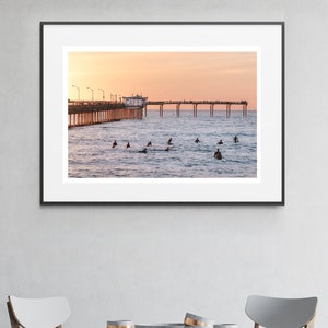 May include: A framed photograph of a pier extending into the ocean at sunset. Surfers are in the water, and people are on the pier. The sky is a gradient of orange and pink.
