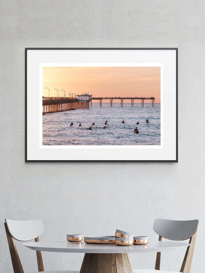 May include: A framed photograph of a pier extending into the ocean at sunset. Surfers are in the water, and people are on the pier. The sky is a gradient of orange and pink.