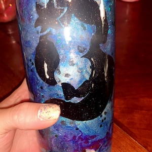 Puede incluir: Un vaso azul y morado con purpurina y una silueta negra de una sirena. El vaso tiene una tapa transparente.