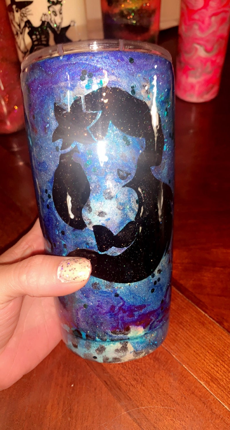 Puede incluir: Un vaso azul y morado con purpurina y una silueta negra de una sirena. El vaso tiene una tapa transparente.