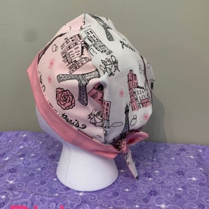 May include: A pink and white surgical scrub hat with a Parisian theme. The hat features a black and white illustration of the Eiffel Tower, buildings, and the word "Paris".