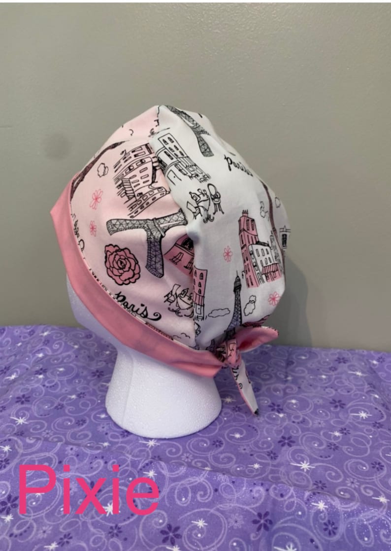 May include: A pink and white surgical scrub hat with a Parisian theme. The hat features a black and white illustration of the Eiffel Tower, buildings, and the word "Paris".
