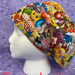 May include: A white scrub hat with a colorful print of various emojis, including smiley faces, hearts, and thumbs up. The hat has a black elastic band.