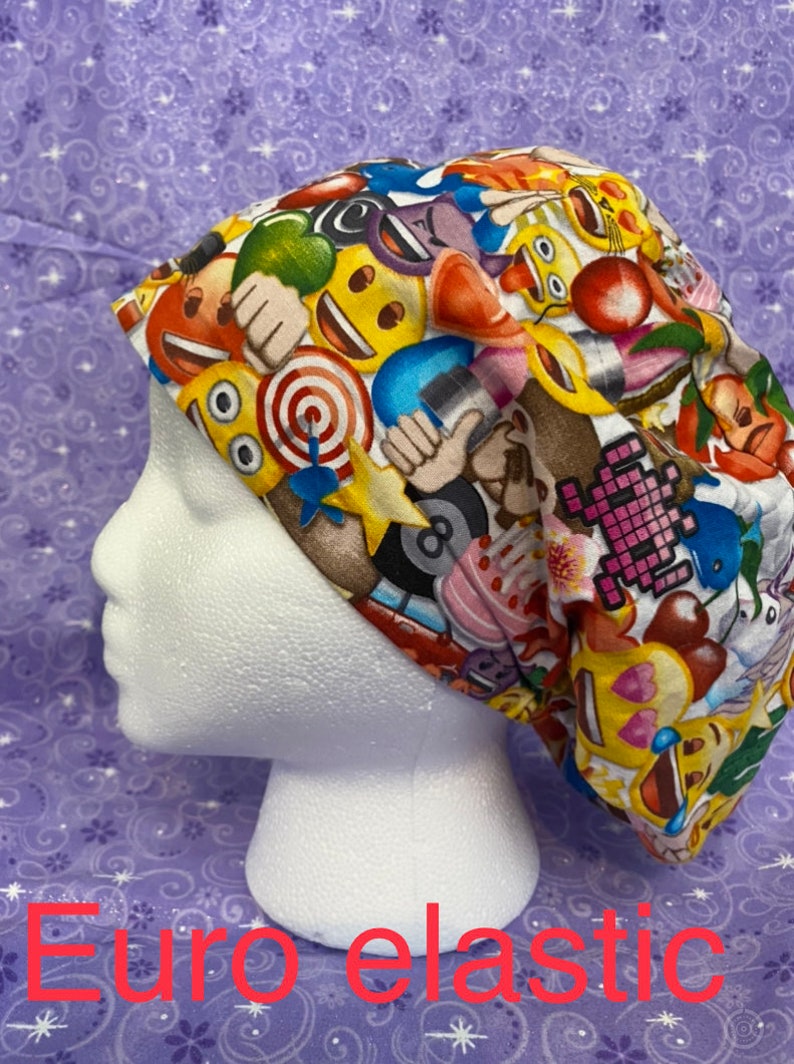 May include: A white scrub hat with a colorful print of various emojis, including smiley faces, hearts, and thumbs up. The hat has a black elastic band.