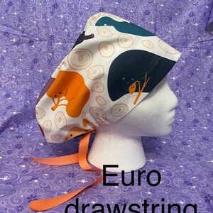 May include: A white scrub hat with a colorful cartoon print of animals and circles. The hat has a peach-colored drawstring.