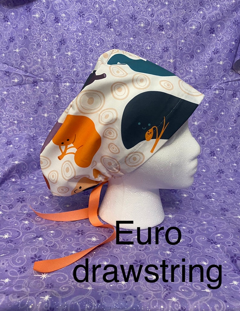 May include: A white scrub hat with a colorful cartoon print of animals and circles. The hat has a peach-colored drawstring.