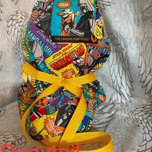 May include: A yellow fabric ponytail holder with a colorful print of comic book style Star Wars Mandalorian characters. The print includes the text "Star Wars The Mandalorian" and "The Legend Continues".