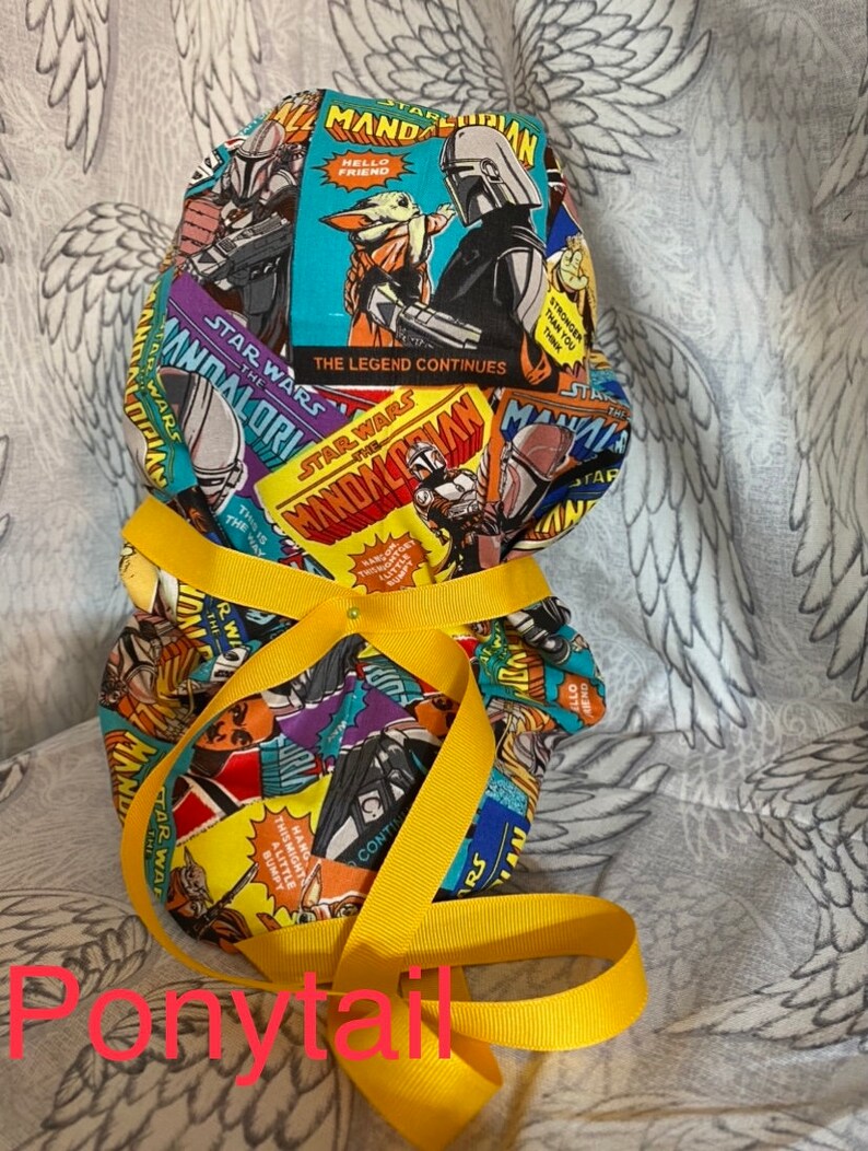 May include: A yellow fabric ponytail holder with a colorful print of comic book style Star Wars Mandalorian characters. The print includes the text "Star Wars The Mandalorian" and "The Legend Continues".