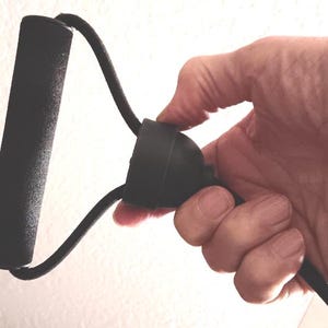 May include: Black resistance band with a foam handle. The band is held in a hand, showing the handle and the connecting cord. The handle is cylindrical and the cord is a thick, flexible material. The background is a plain white wall.