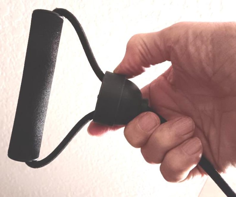 May include: Black resistance band with a foam handle. The band is held in a hand, showing the handle and the connecting cord. The handle is cylindrical and the cord is a thick, flexible material. The background is a plain white wall.