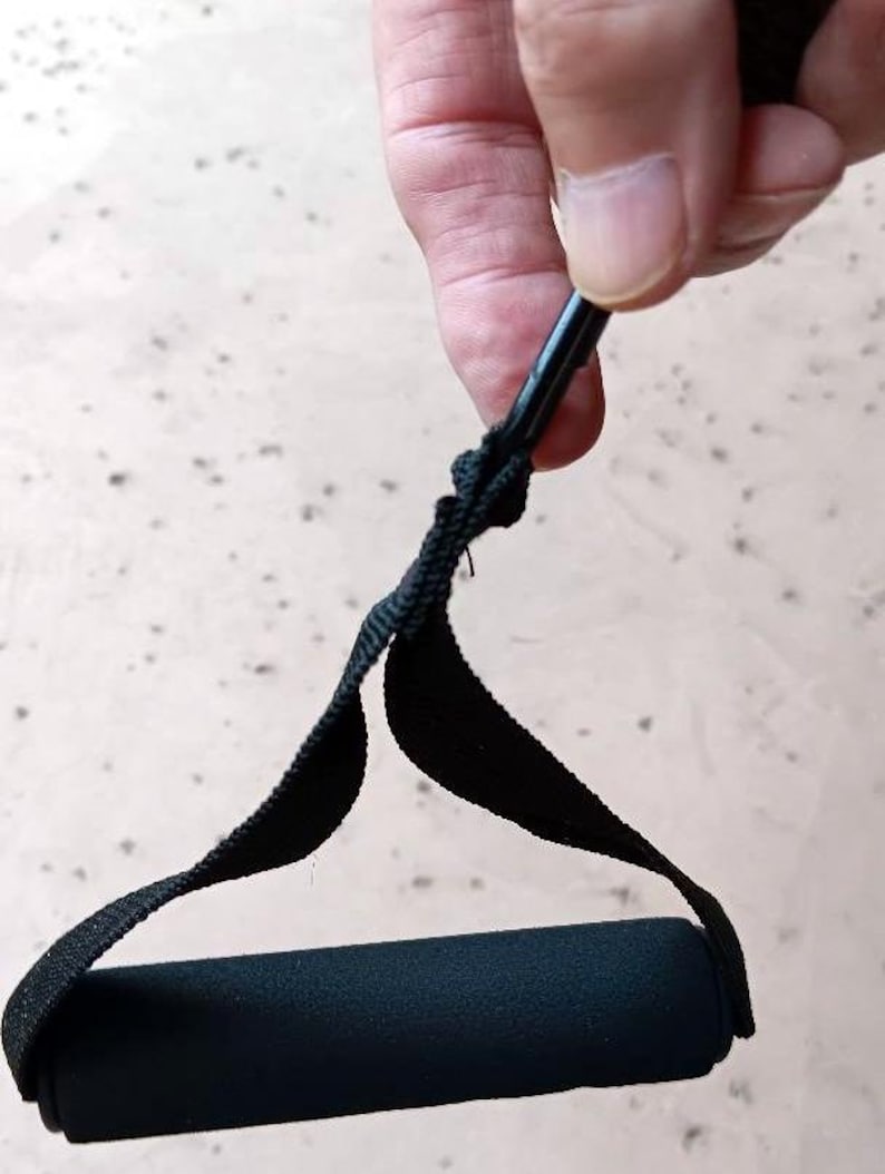 May include: Black exercise resistance band handle with a black foam grip. The handle is attached to a black strap and a black metal rod. The handle is held by a person.