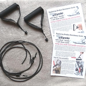 May include: A set of black exercise equipment replacement parts, including two handles, a long cord, and two instruction sheets. The text on the sheets reads "Replacing Broken Resistance Cords" and "Slim Cycle".