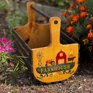 May include: A wooden carrier with a dark handle and black slatted sides. The front panel features a natural wood finish with a painted graphic of a windmill, a red tractor, a red barn, and a black cow. The word FARMHOUSE is printed in gold.