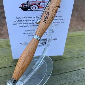 May include: A wooden pen with a silver clip and tip, displayed on a clear stand. The pen is engraved with the "Packard" logo. A card behind the pen states the pen is made from wood salvaged from the Packard Automotive Plant.