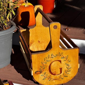 May include: A wooden harvest basket with a handle and a decorative front panel. The panel features a green and red wreath design around a large letter "G". The basket is made of light-colored wood and is designed for carrying items.