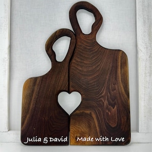 Puede incluir: Dos tablas de cortar de madera, unidas para formar un corazón. Las tablas son de color marrón oscuro con vetas naturales. Una tabla está inscrita con "Julia & David", la otra con "Made with Love".