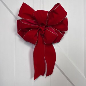 May include: A large, red velvet bow with a gold trim. The bow is tied with a long, flowing tail and is hanging on a white door.