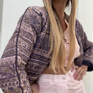 Puede incluir: Una mujer con una chaqueta acolchada con estampado morado y marrón, un top de crochet rosa y pantalones rosa claro.