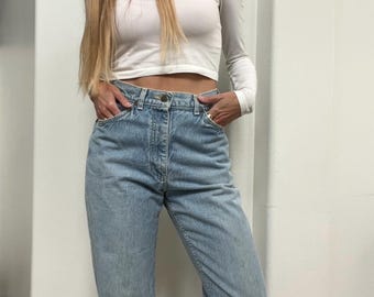 May include: A woman in a white off-the-shoulder long-sleeve crop top and light blue jeans. The jeans are high-waisted and cuffed. She is also wearing pink ankle boots.