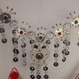 Pode incluir: Colar de prata com um pingente de coração e pingentes de moedas. O pingente de coração é decorado com filigrana e pedras preciosas vermelhas e verdes. Os pingentes de moedas também são decorados com pedras preciosas vermelhas e verdes.