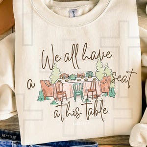 May include: A beige sweatshirt with a brown graphic of a table with chairs and a teapot. The text "We all have a seat at his table Luke 14:14-17" is printed on the sweatshirt.