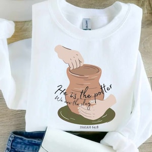 May include: White sweatshirt featuring a graphic of a potter's hands shaping a brown clay pot. The text "He is the potter, We are the clay" is written in black script, with "Isaiah 64:8" below.