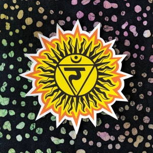May include: A yellow sticker with a sun and a symbol inside. The symbol is a triangle with a circle at the top and a line going down the middle. The sticker is on a black background with white and yellow dots.