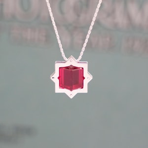 May include: A silver necklace with a geometric pendant. The pendant features a vibrant red gemstone set within a star-shaped frame. The chain is a delicate, twisted design.