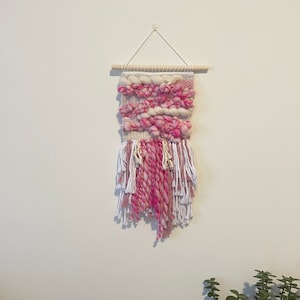 Op de afbeelding: Een roze en witte geweven wandhanger met franjes. De hanger is gemaakt van dik garen en heeft een houten lat aan de bovenkant.