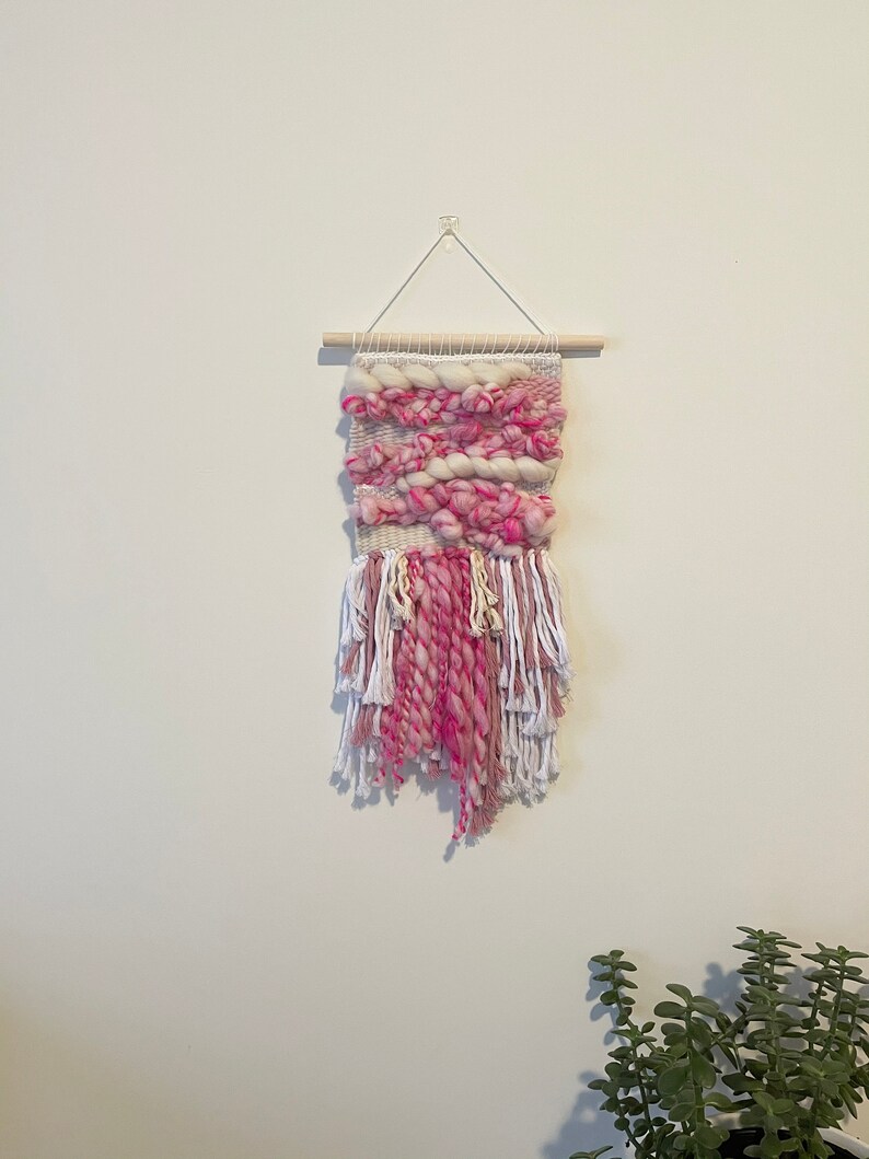 Op de afbeelding: Een roze en witte geweven wandhanger met franjes. De hanger is gemaakt van dik garen en heeft een houten lat aan de bovenkant.