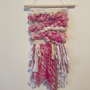 Op de afbeelding: Een roze en witte geweven wandhanger met franjes. De hanger is gemaakt met verschillende texturen en kleuren, waaronder roze, wit en cr&egrave;me.
