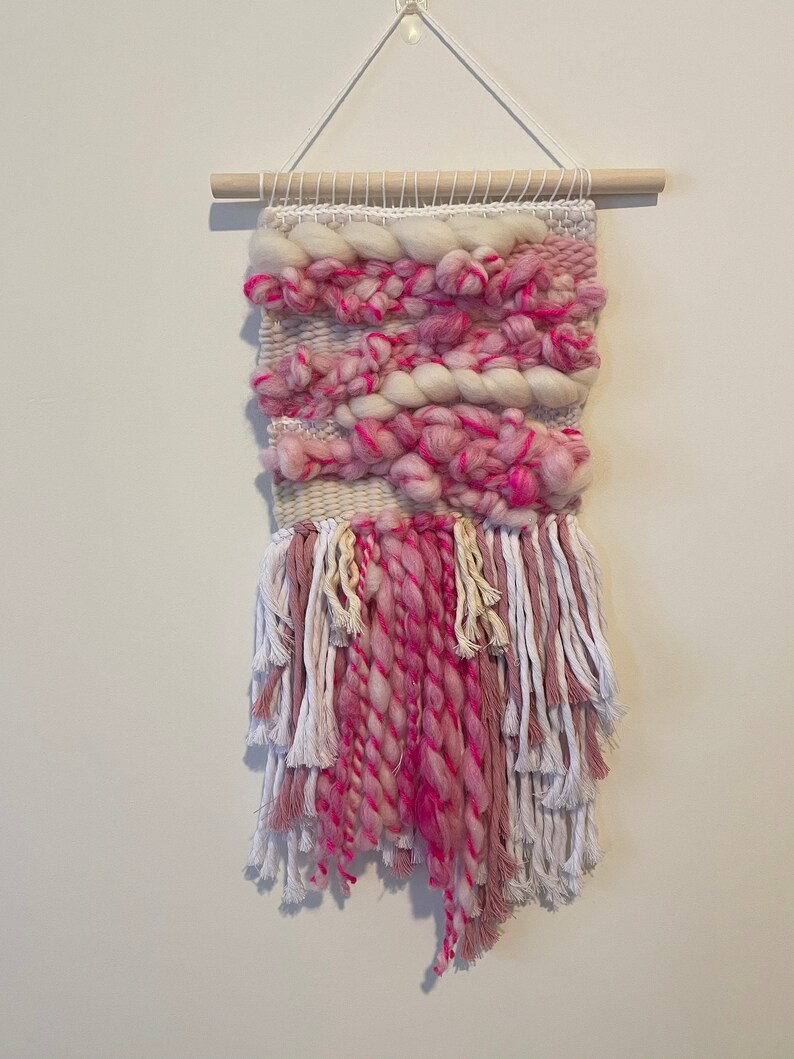 Op de afbeelding: Een roze en witte geweven wandhanger met franjes. De hanger is gemaakt met verschillende texturen en kleuren, waaronder roze, wit en cr&egrave;me.