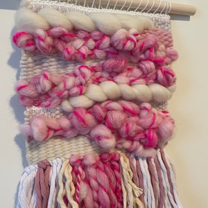Op de afbeelding: Een roze en witte geweven wandhanger met franjes. De hanger is gemaakt met verschillende texturen en kleuren, waaronder roze, wit en cr&egrave;me.