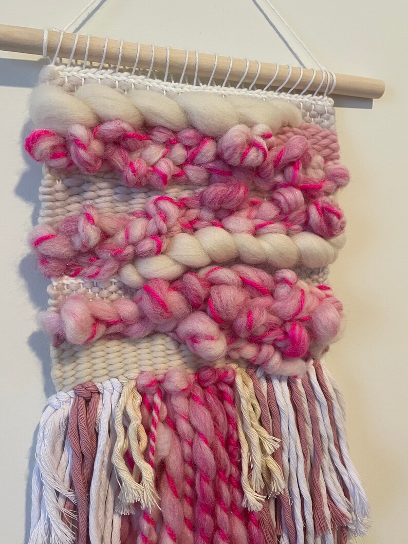 Op de afbeelding: Een roze en witte geweven wandhanger met franjes. De hanger is gemaakt met verschillende texturen en kleuren, waaronder roze, wit en cr&egrave;me.